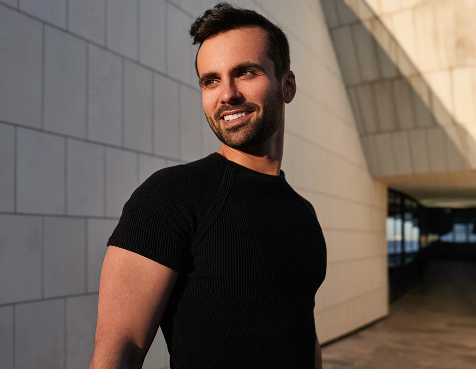 A man with a trimmed beard and dark hair, wearing a fitted black shirt, smiles confidently as he stands outside in the sunlight, with modern architecture in the background - Gynecomastia Procedure in Houston, TX